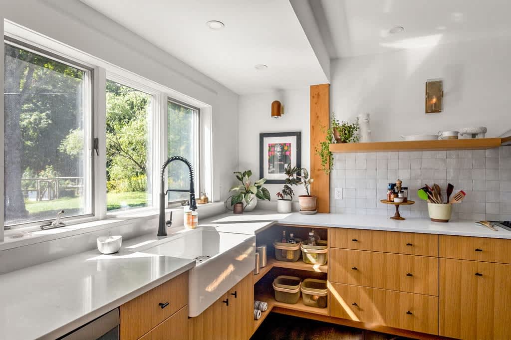 A modern kitchen features light wood cabinetry, white countertops, a large farmhouse sink with a black faucet under a wide window, and various potted plants adding a touch of greenery, with dark hardwood floors.