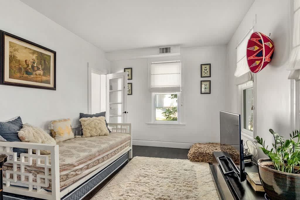 A bright room features a white daybed with patterned bedding and plush pillows, a light-colored shaggy rug, and a wall-mounted TV, with natural light streaming through a window and dark hardwood floors.