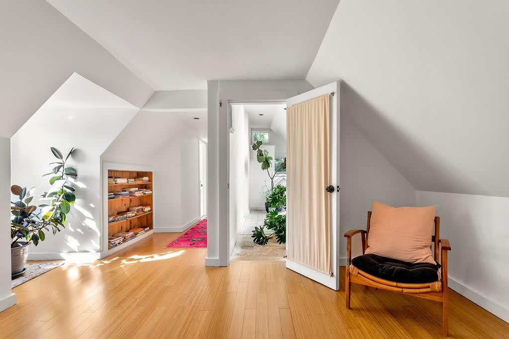 Bright hallway with light wood floors, white walls, and sloped ceilings, featuring built-in wooden shelves with books, several potted plants, and an open doorway leading to another room.