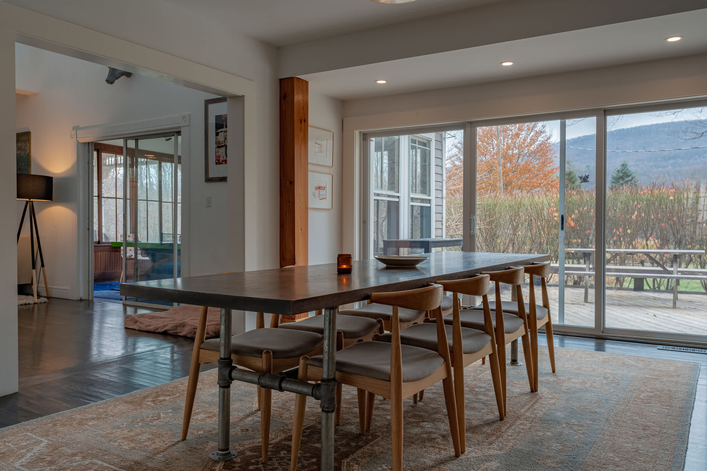 Spacious dining room with a long wooden table and six chairs, set on a patterned rug, featuring large sliding glass doors that open to an outdoor deck and reveal a wooded landscape.