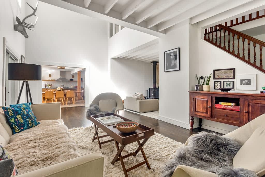 A spacious living room with dark hardwood floors and white exposed beams features multiple seating areas, including a light sofa with faux fur throws, a wooden coffee table, and a dark wooden staircase leading upstairs.