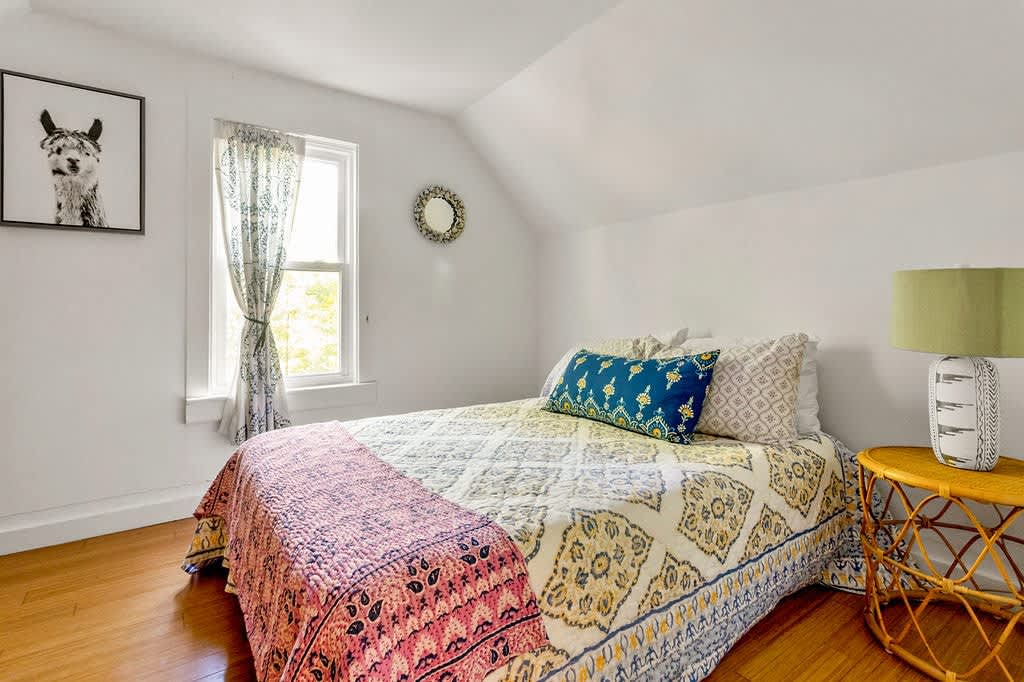 Bright bedroom with a patterned bedspread, a rattan nightstand with a green lamp, and a window with sheer curtains, featuring a framed llama art print and a round mirror.