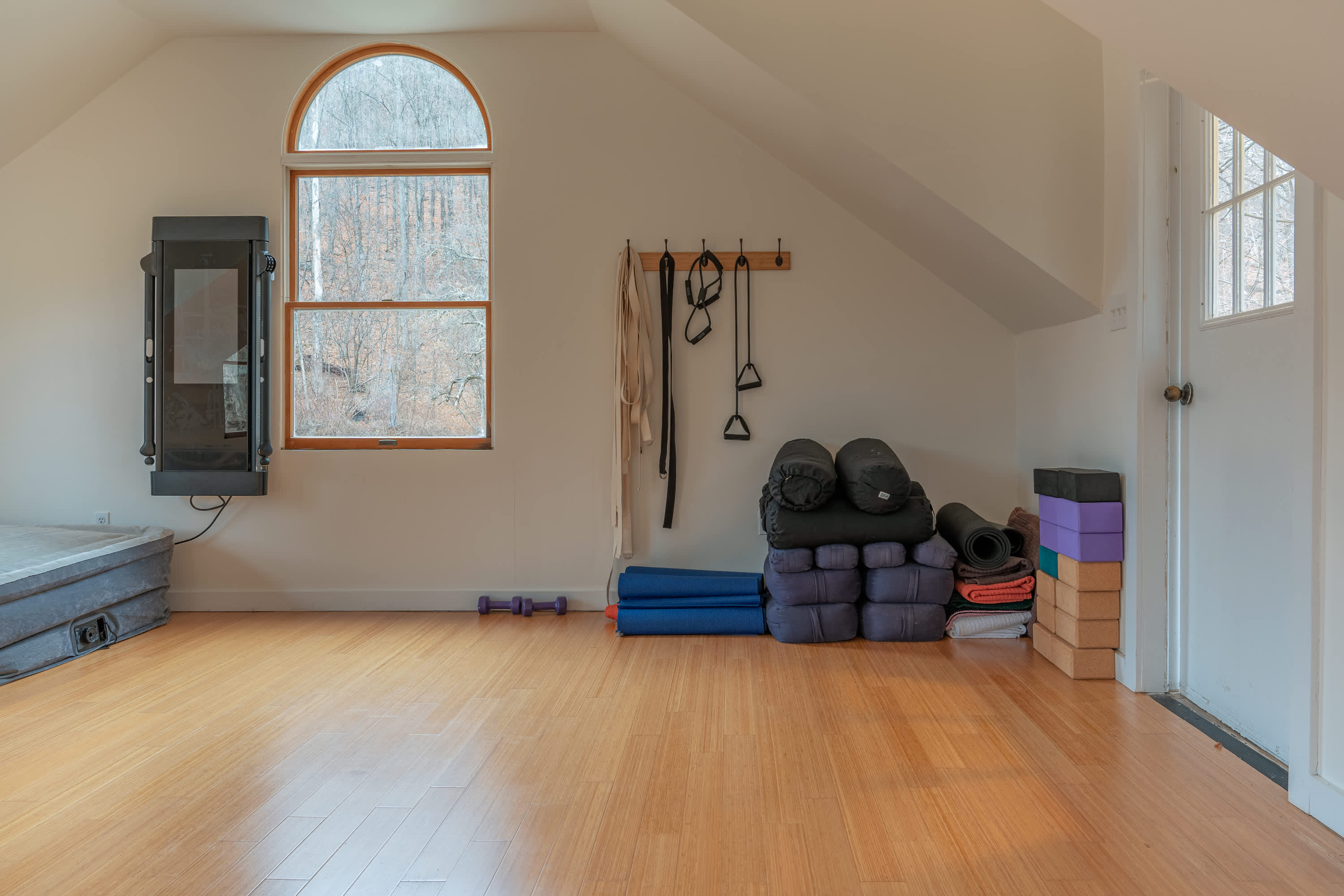 Spacious yoga studio with light wood floors and white walls, featuring a large arched window, various yoga mats, blocks, and straps, and a wall-mounted exercise machine.