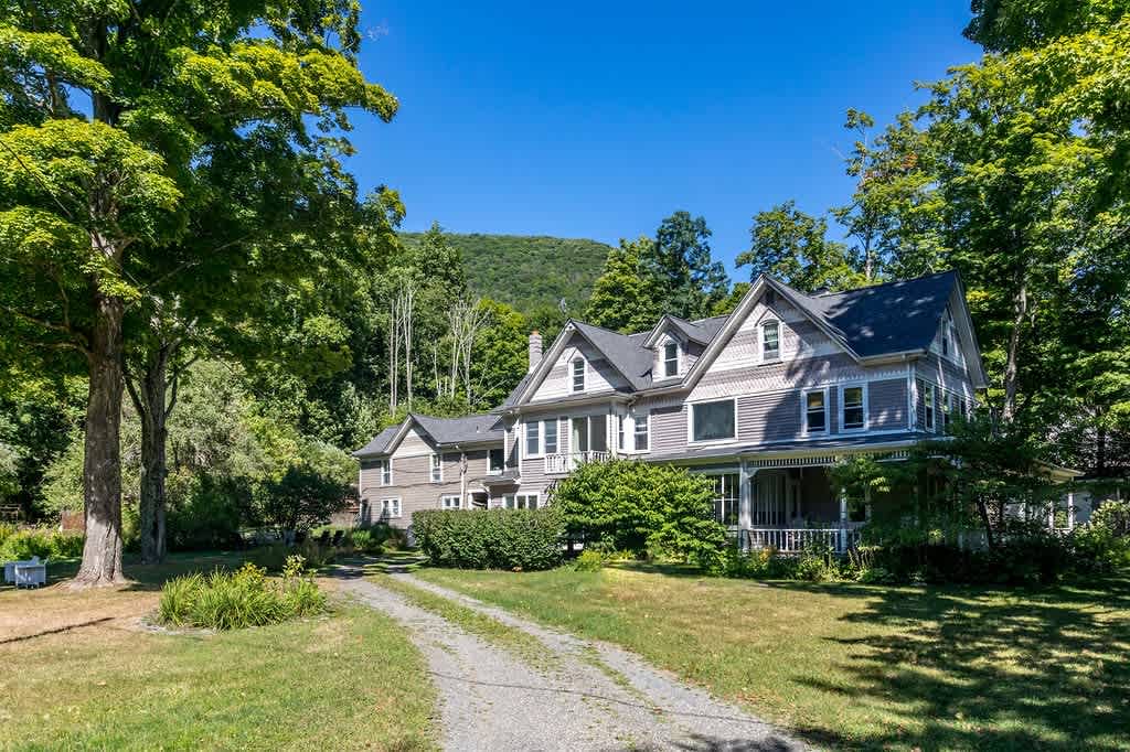 The grand front exterior of a multi-story house with gray siding and white trim, featuring multiple gables and a covered porch, nestled amidst mature green trees with a mountain backdrop.