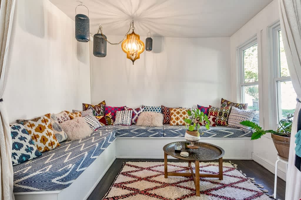 A vibrant seating nook features a built-in L-shaped bench with patterned cushions and numerous colorful throw pillows, centered around a round metal coffee table, under unique hanging lanterns and a patterned rug.