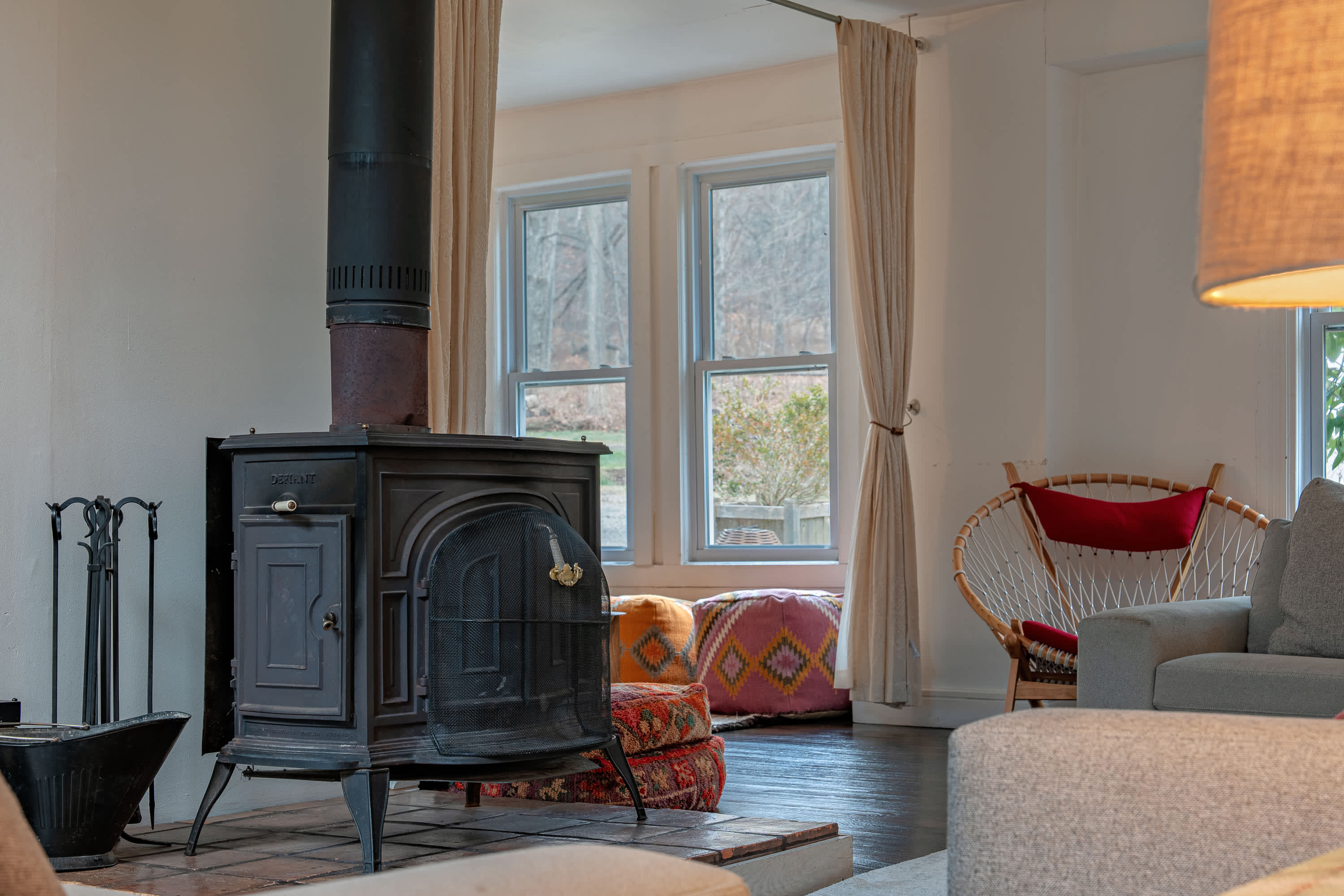 A detailed view of a black cast-iron wood-burning stove with a mesh door, set on a tiled hearth, flanked by a metal log holder and tools, with a window revealing outdoor greenery in the background.