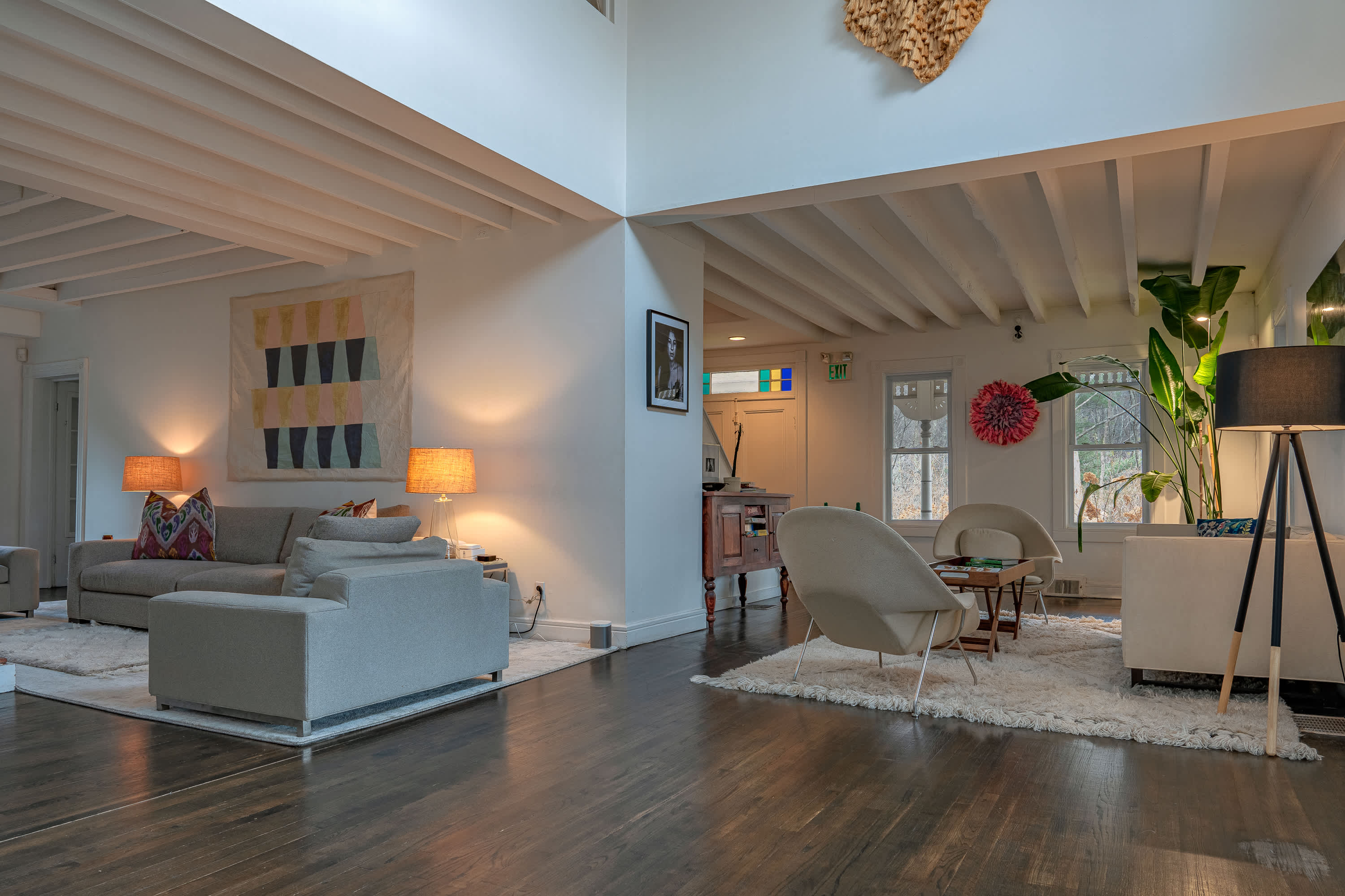 A large, open-concept living space with dark hardwood floors and white exposed beams features various seating arrangements, including a light sofa and armchairs, illuminated by lamps and natural light from tall windows.