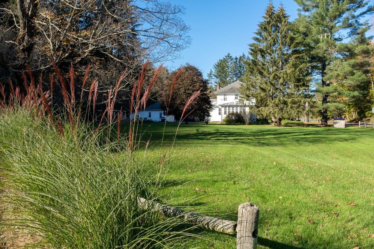 A wide shot of a spacious green yard with tall ornamental grasses in the foreground and a rustic wooden fence post, leading to a distant white house nestled among mature trees under a clear blue sky.