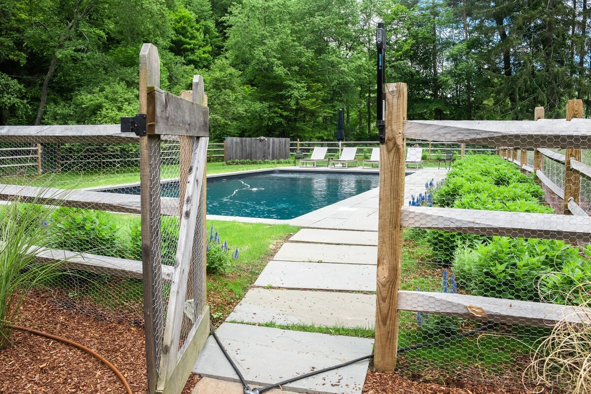 A clear blue swimming pool is visible through an open wooden gate with chicken wire fencing, leading to a stone path surrounded by lush green foliage and tall trees in the background.
