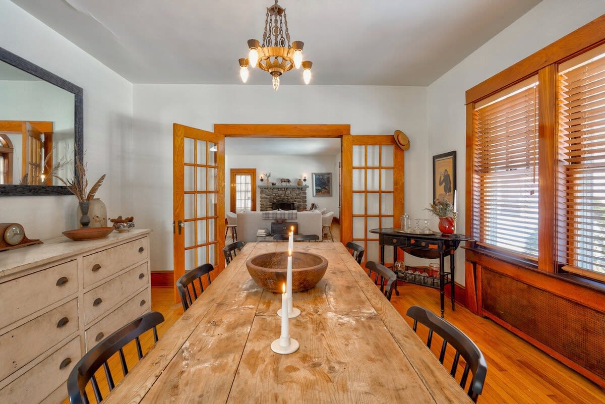 A rustic wooden dining table with candles and a large wooden bowl is centered in a dining room with hardwood floors, white walls, and a vintage chandelier, with French doors leading to another living space.
