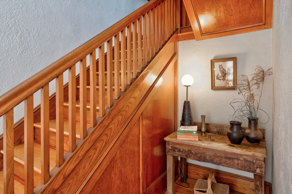 Inviting foyer featuring a polished wooden staircase and banister, with a rustic console table adorned with books, vases, and a modern lamp, set against a light wall.