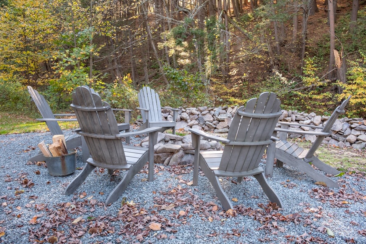 A cozy outdoor fire pit area features several grey Adirondack chairs arranged in a circle around a stone fire pit, set on a gravel ground scattered with autumn leaves, backed by a dense forest.