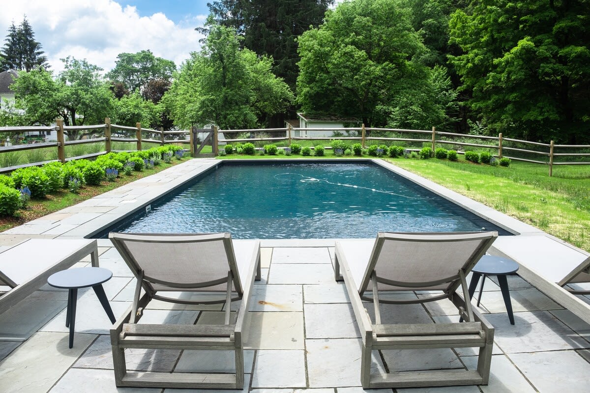 Rectangular outdoor swimming pool surrounded by light-colored stone paving, with two modern lounge chairs and side tables. A wooden fence borders a lush green lawn and mature trees under a partly cloudy sky.