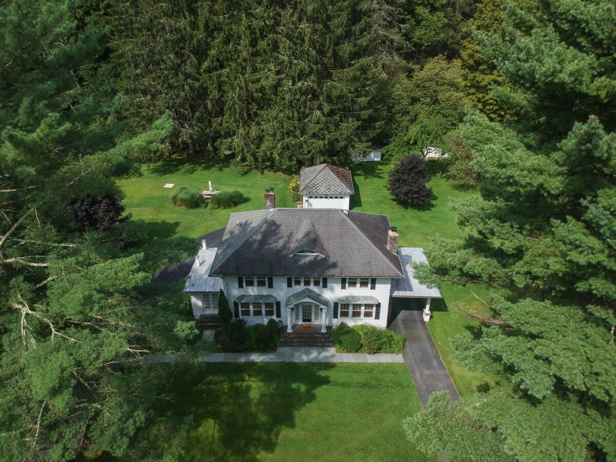 An aerial view showcases a large white house with a dark roof, surrounded by expansive green lawns and dense evergreen trees, with a smaller outbuilding visible in the distance.