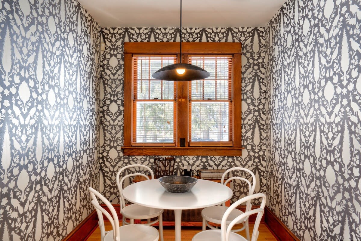 Cozy dining nook featuring bold black and white floral wallpaper, a round white table with four matching chairs, and a modern black pendant light hanging above. A large wooden window with blinds provides natural light.