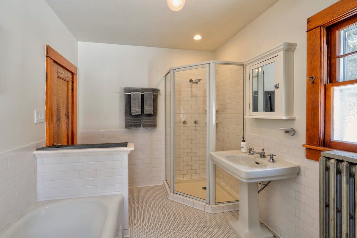 Classic white tiled bathroom featuring a corner glass shower, a pedestal sink, and a built-in bathtub, with a window providing natural light and a vintage radiator.