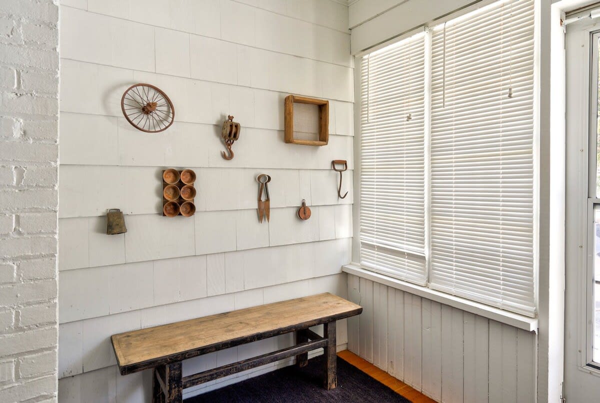 Bright enclosed porch or mudroom with white shiplap walls adorned with rustic metal and wooden decor, featuring a long wooden bench and a window with blinds, next to a brick wall.