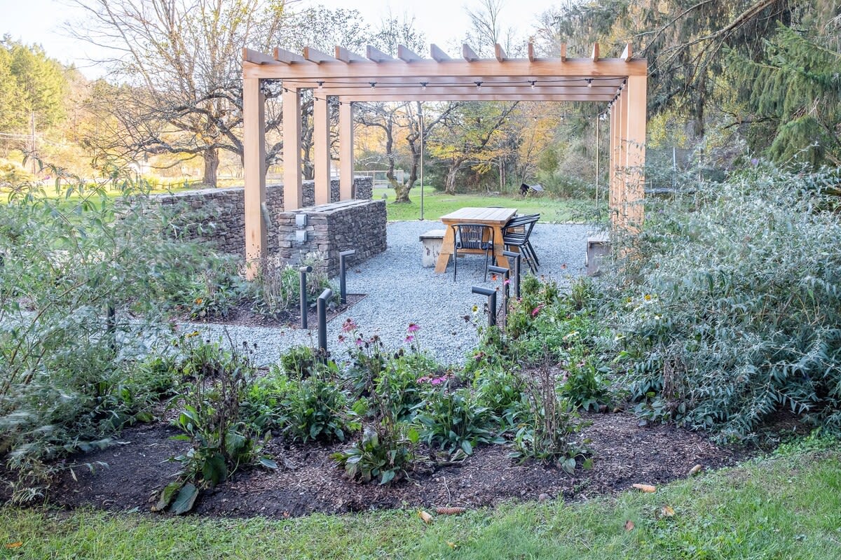 An outdoor dining area under a light wood pergola, with a long wooden table and chairs, is surrounded by a gravel path and garden beds with various plants, backed by mature trees.