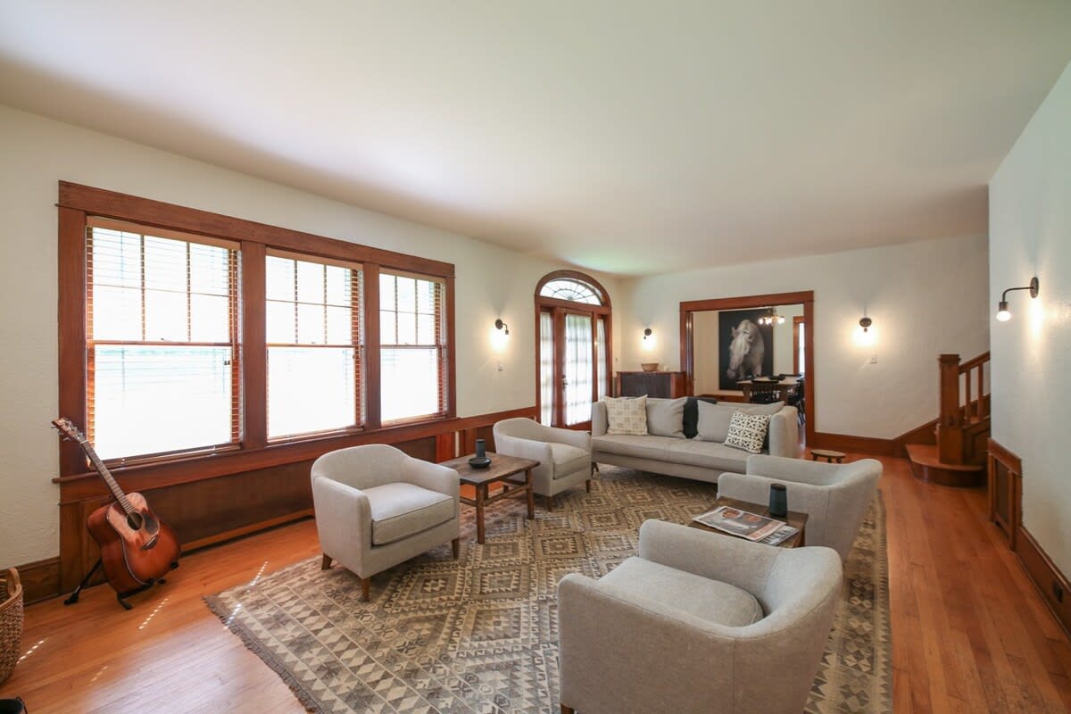 Spacious living room featuring light gray armchairs and a sofa, a patterned area rug on hardwood floors, and large windows with wooden blinds. Warm wooden trim accents the windows and doorways, leading to other rooms.