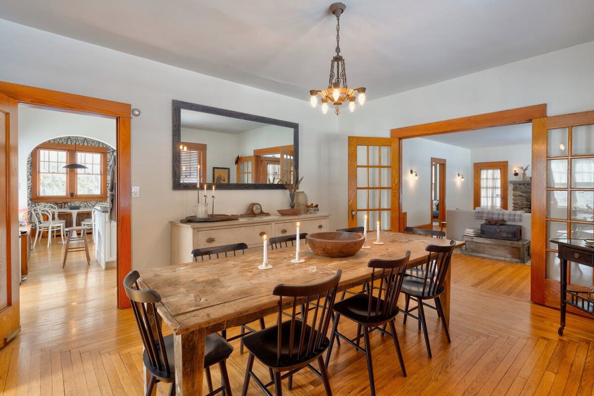 Formal dining room with a long rustic wooden table, black spindle-back chairs, and a vintage chandelier. Hardwood floors with a parquet pattern extend throughout, and a large mirror hangs above a buffet cabinet.