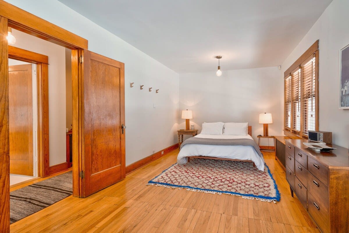 Spacious bedroom with a white bed, two wooden nightstands with lamps, a long wooden dresser, and a colorful patterned area rug on light hardwood floors, with wooden doors.