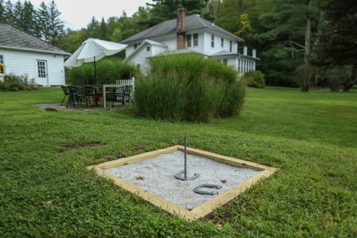 A horseshoe pit with two horseshoes and a stake is set in a well-maintained green lawn, with a white house and outdoor seating area partially visible in the background.