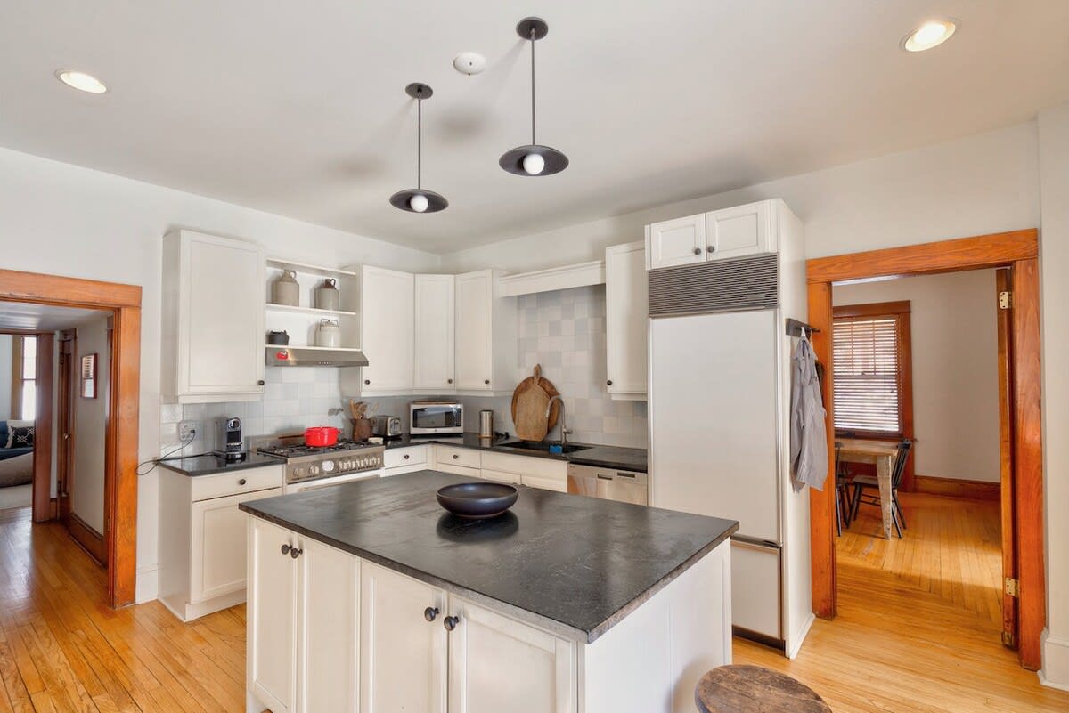 A spacious kitchen features white shaker cabinets, a large island with a dark countertop, stainless steel appliances, and hardwood floors, with open doorways leading to other rooms.