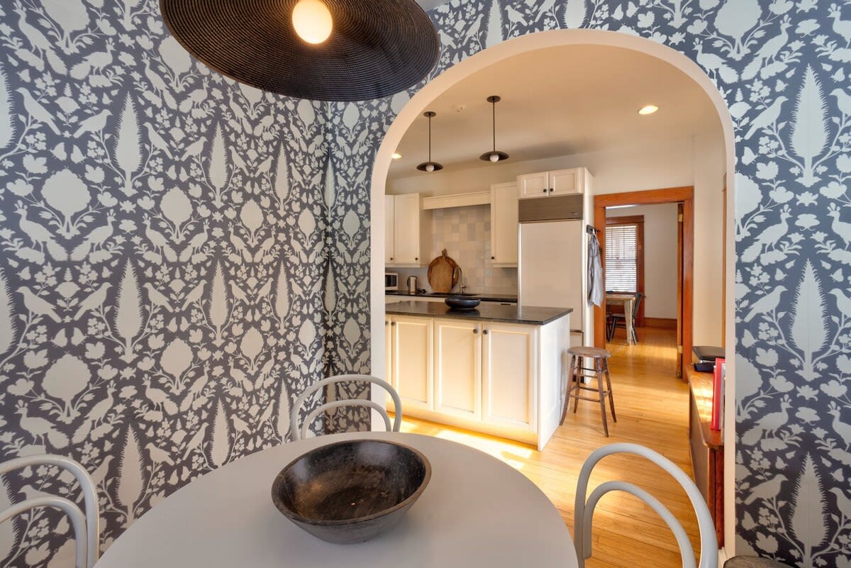 Dining nook with striking black and white floral wallpaper, a round white table, and an arched doorway leading into a bright kitchen with white cabinets and dark countertops.