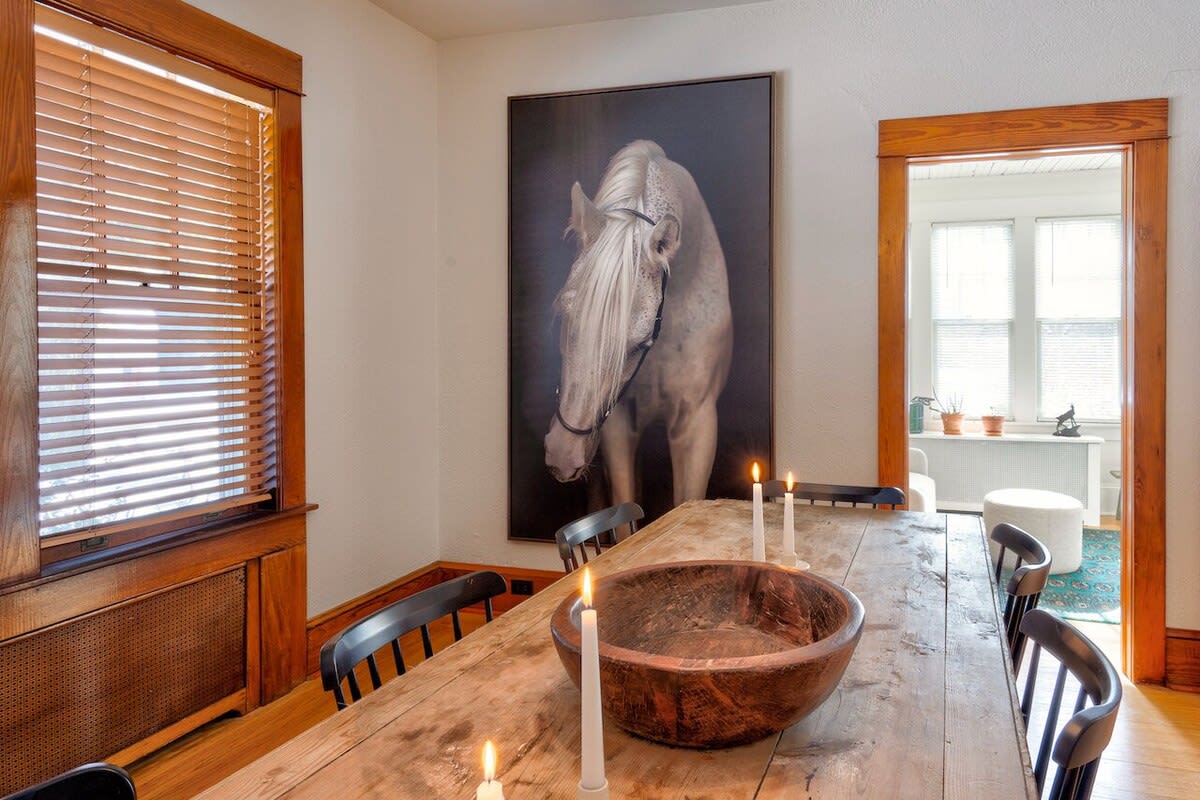 A rustic wooden dining table is positioned in a room with warm hardwood floors and white walls, featuring a large framed horse portrait, wooden blinds on the windows, and an open doorway to an adjacent room.