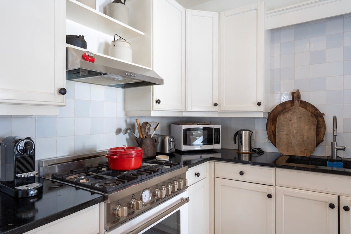 Modern kitchen featuring white shaker-style cabinets, black countertops, and a light blue tiled backsplash. Stainless steel appliances include a gas range with a red pot, a microwave, and a coffee machine.