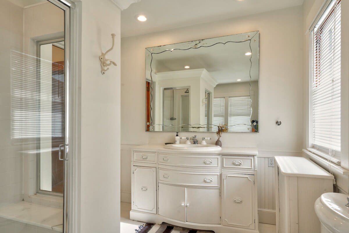 Elegant white bathroom featuring a large ornate mirror above a curved vanity with a built-in sink, and a glass-enclosed shower visible to the left, complemented by wainscoting.