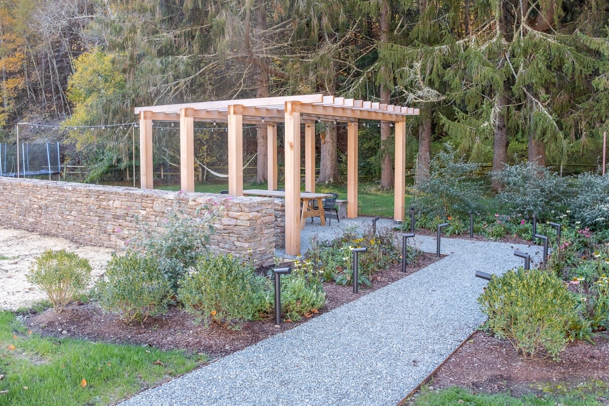 A wooden pergola covers an outdoor seating area with a table, adjacent to a low stone wall and a gravel path lined with garden lights and plants, with a trampoline and forest in the background.