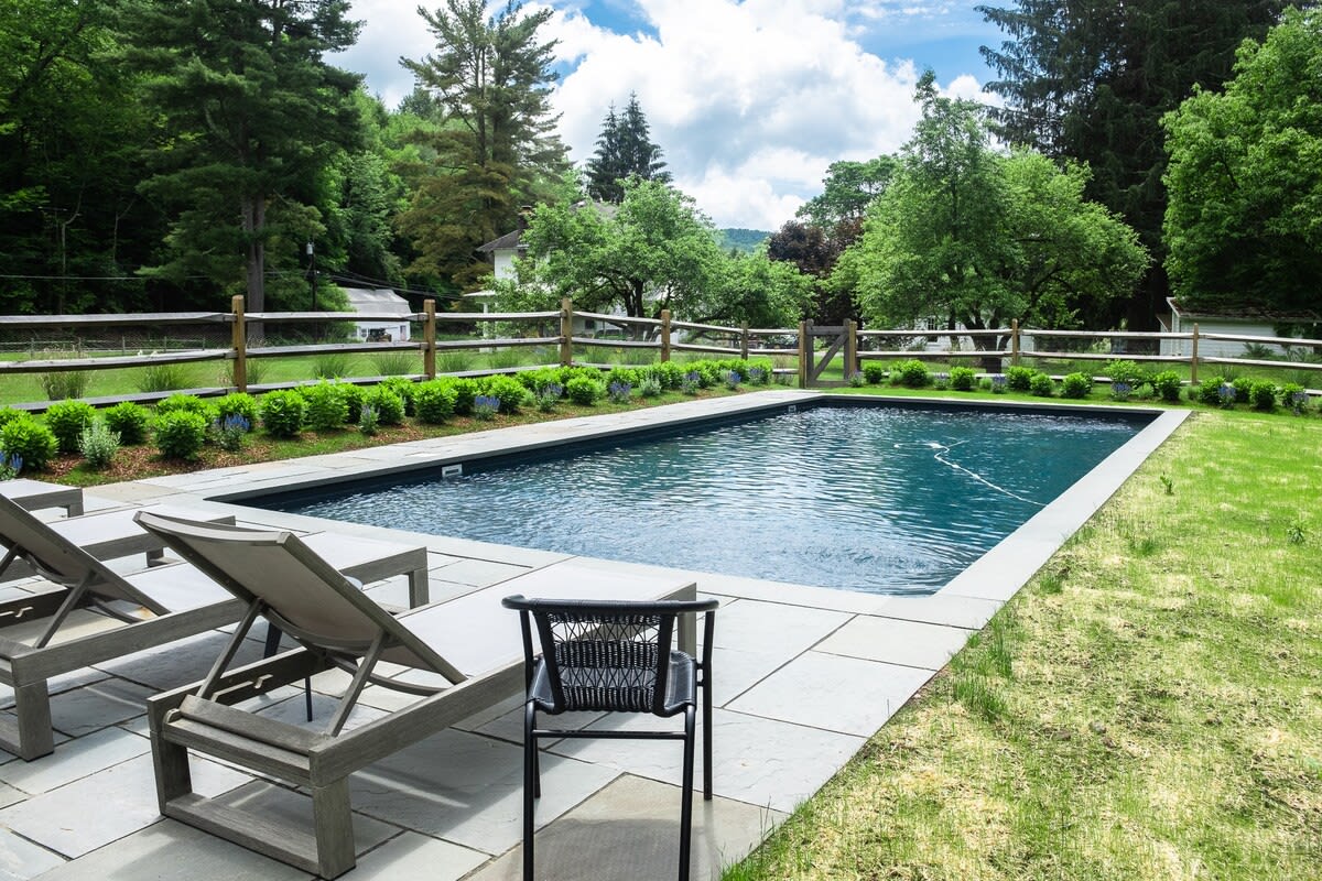 A rectangular swimming pool with dark blue water is bordered by a light-colored stone patio, furnished with several modern lounge chairs and a black outdoor chair, overlooking a manicured lawn and a distant wooden fence with trees.