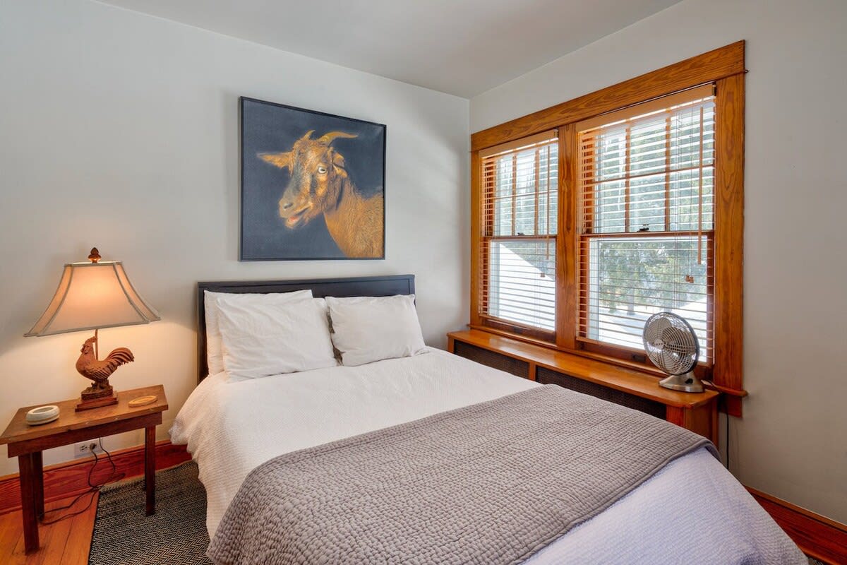 Cozy bedroom with a white bed, grey throw, and a unique goat portrait above the headboard, featuring a large window with wooden blinds and trim, and a small nightstand with a lamp.