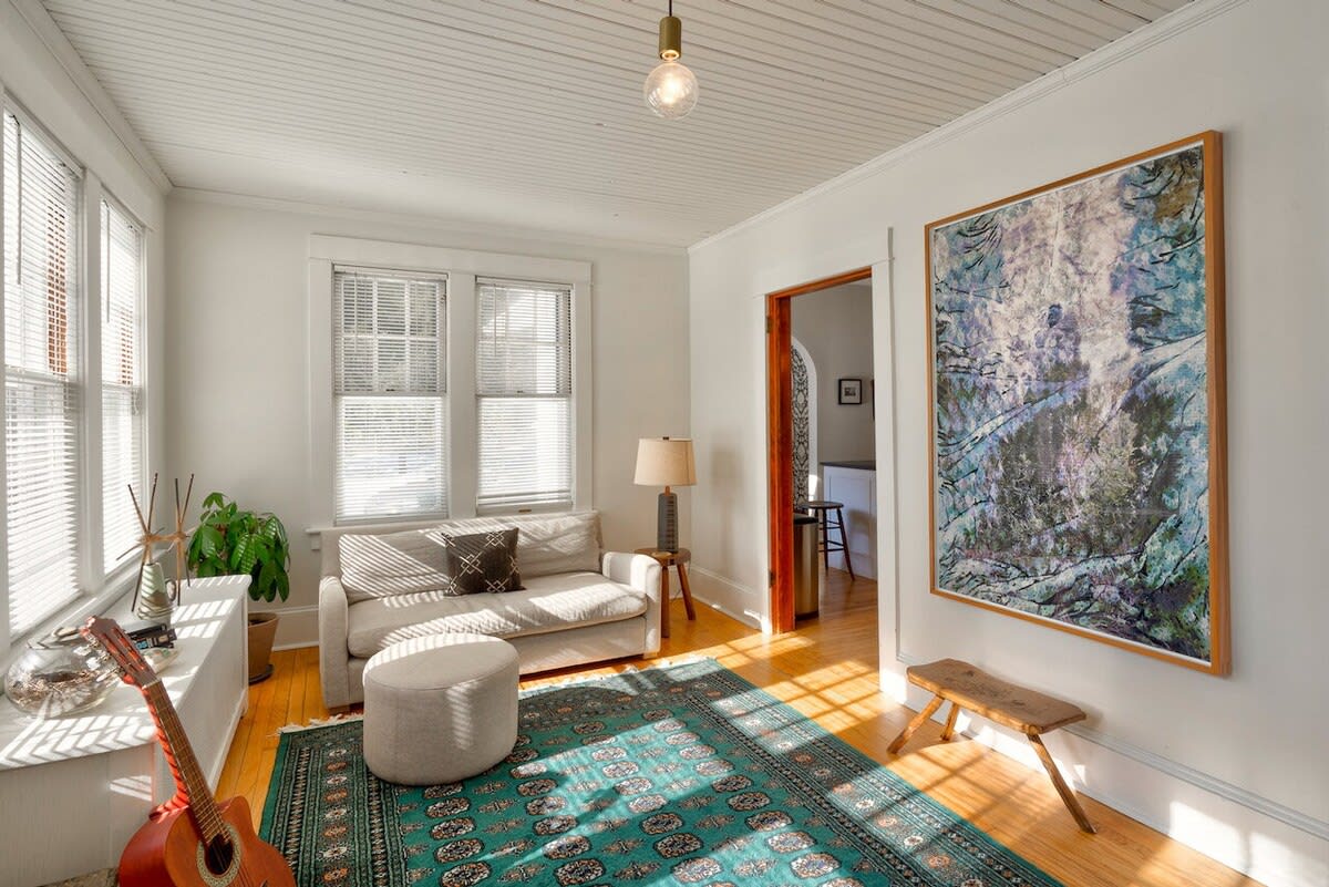 A bright sunlit sitting area features a small sofa, an ottoman, and a guitar, set on a patterned rug over hardwood floors, with multiple windows providing natural light and a large abstract painting on the wall.