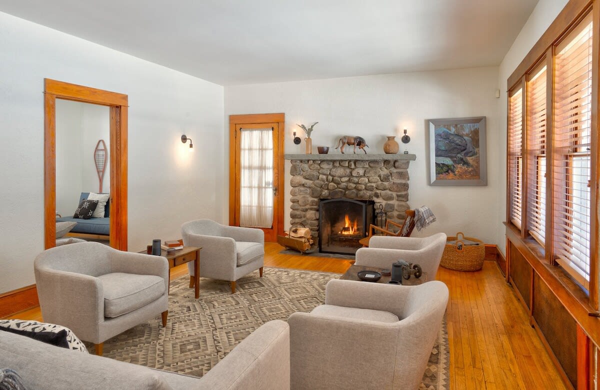 Inviting living room with a rustic stone fireplace, light-colored armchairs arranged around a patterned rug, and warm hardwood floors. Wooden trim frames the windows and doorways, adding a cozy cabin feel.