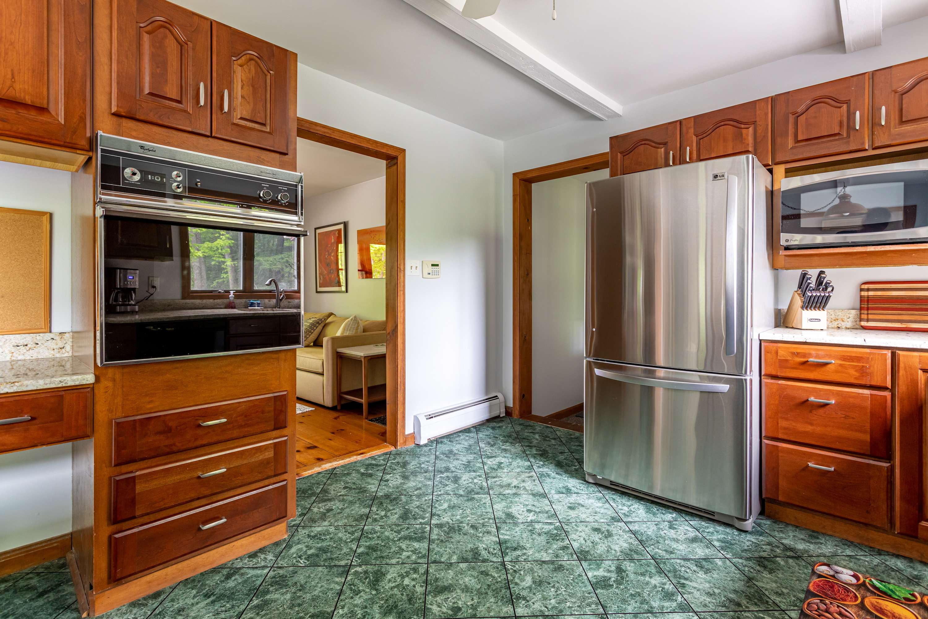 A kitchen with dark wood cabinetry and green tiled floors features a stainless steel refrigerator, a microwave, and a built-in oven, with an open doorway leading to the living area.