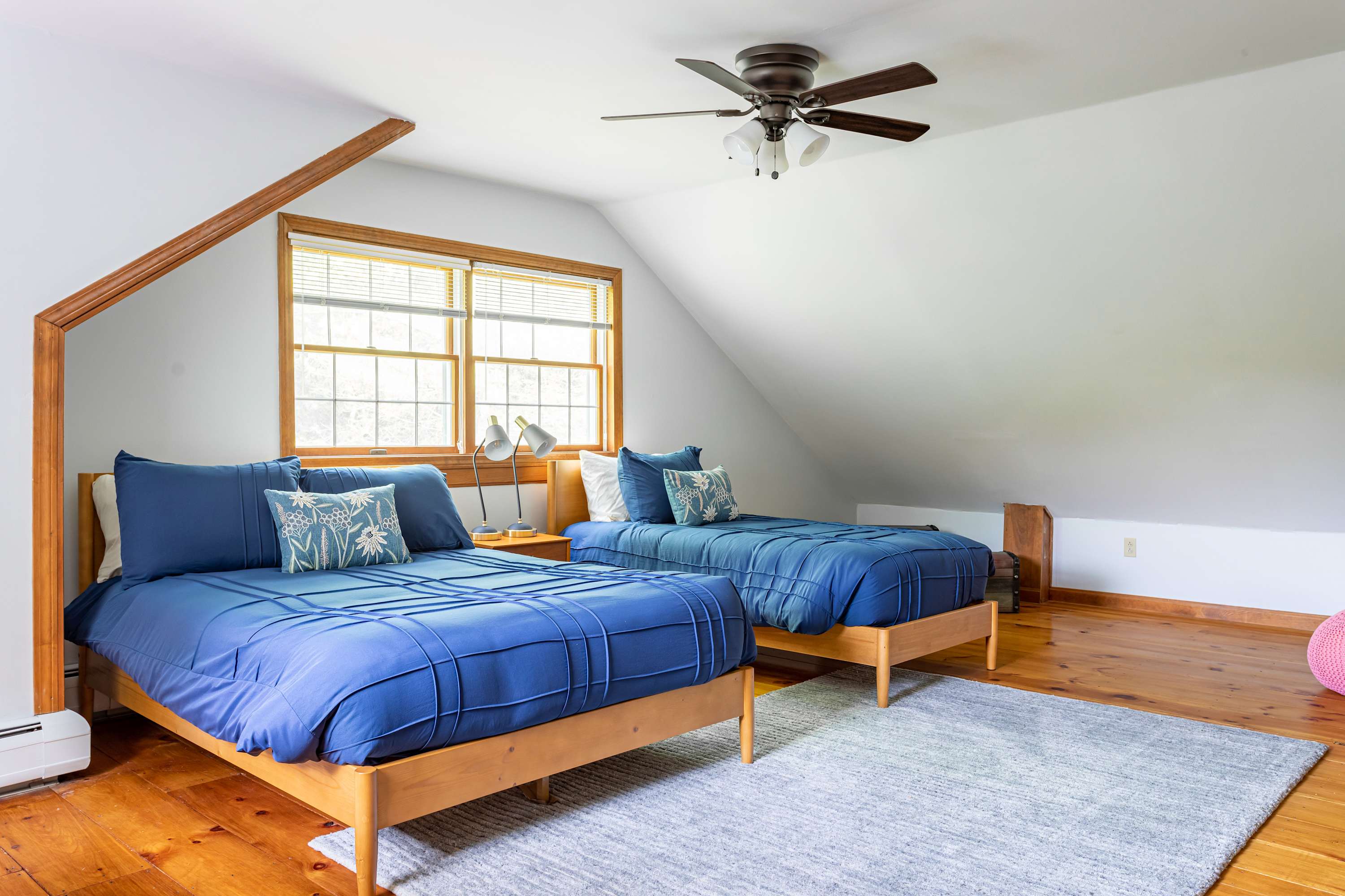 Another view of the bedroom with two twin beds, blue bedding, and light wood floors, showcasing a large window with blinds and a ceiling fan.