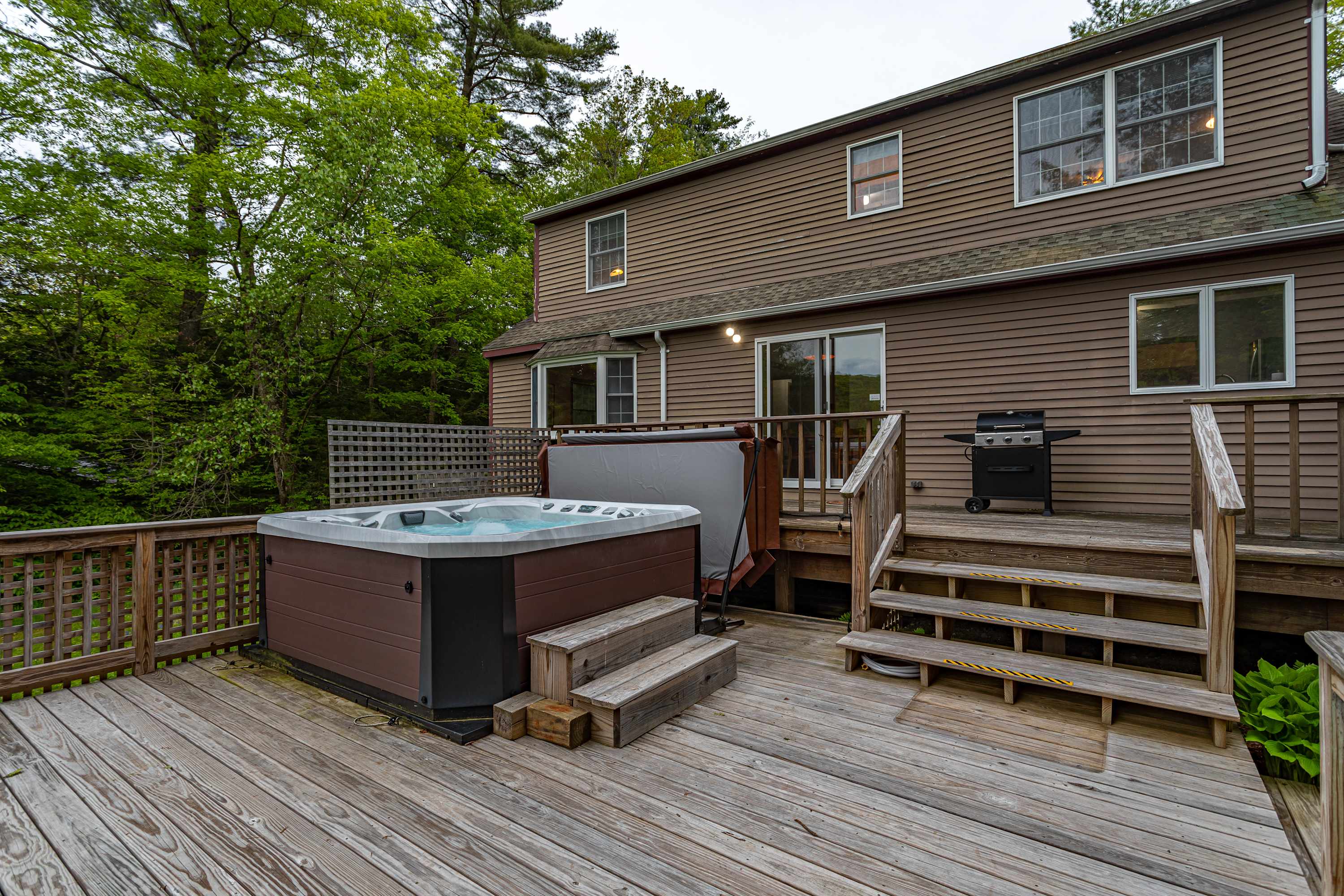 A spacious wooden deck features a large hot tub with a gray cover, a BBQ grill, and wooden steps leading up to the brown-sided house with multiple windows and sliding glass doors, surrounded by lush green trees.