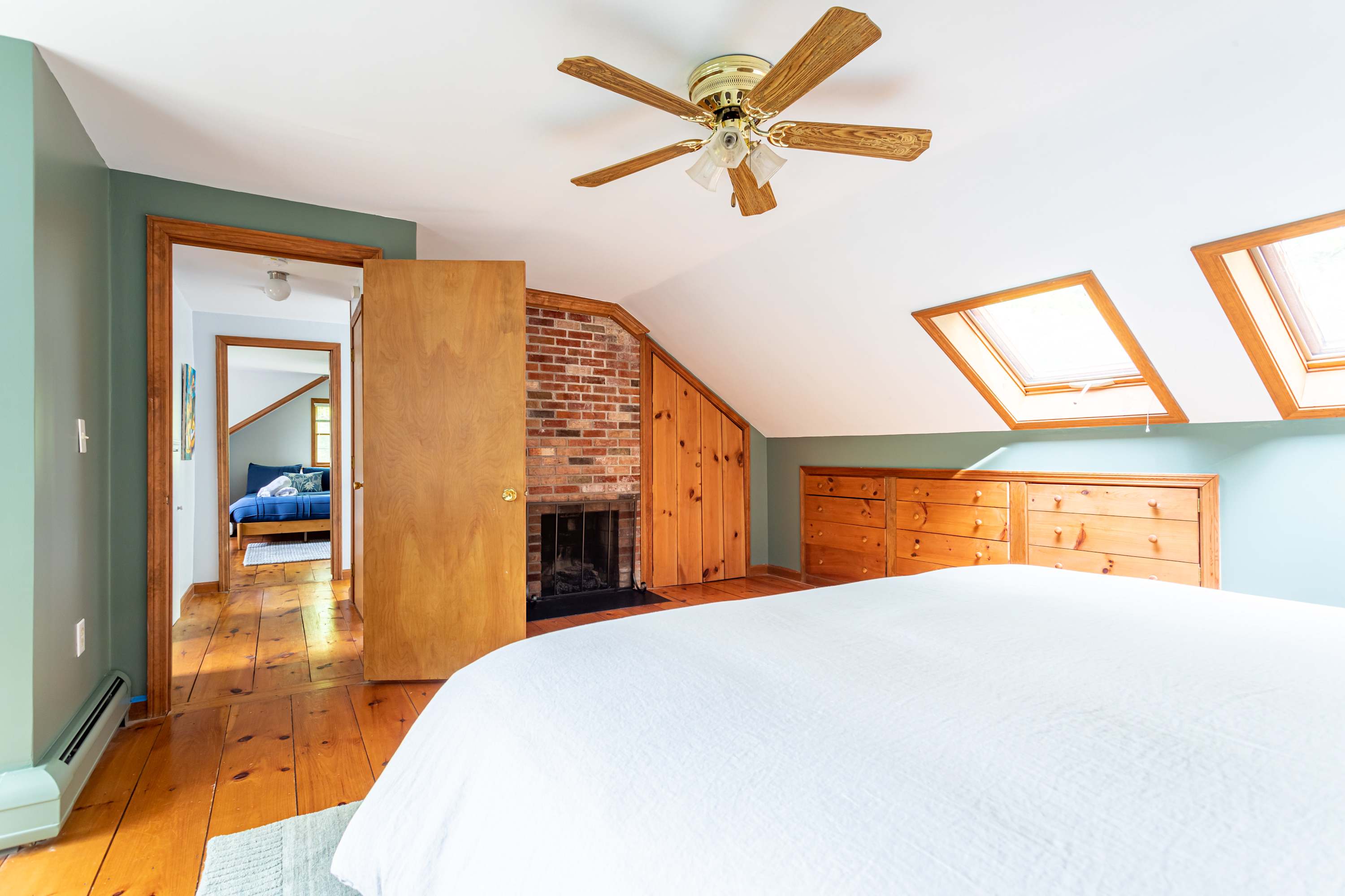 Bright attic bedroom with a white bed, light wood floors, a brick fireplace, and two skylights, featuring a ceiling fan and a doorway leading to another bedroom.
