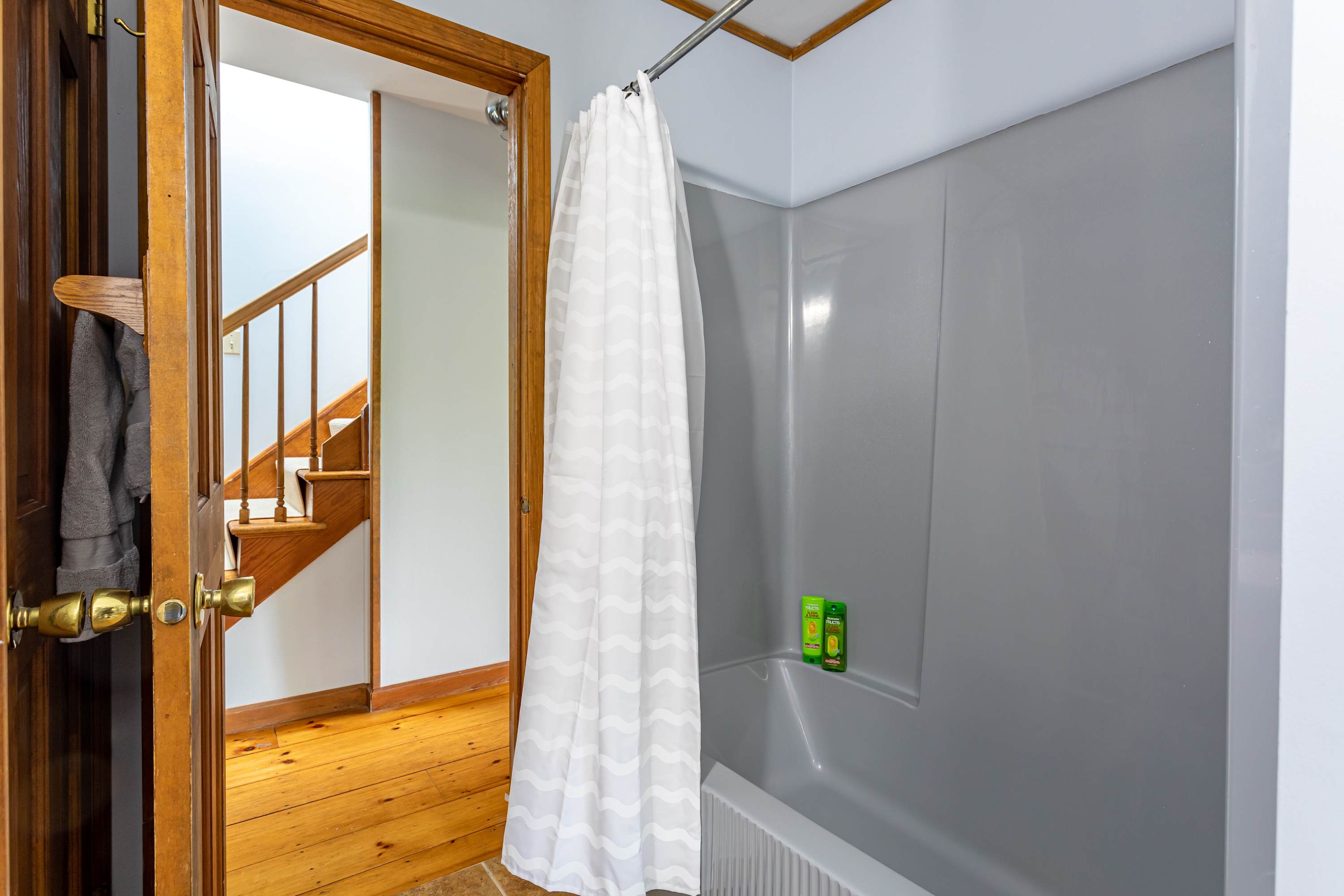 A view inside bathroom-1, showcasing a shower/tub combination with a white shower curtain and a gray surround. The light brown tiled floor extends throughout, and a wooden doorway leads to an adjacent room, maintaining a cohesive design.