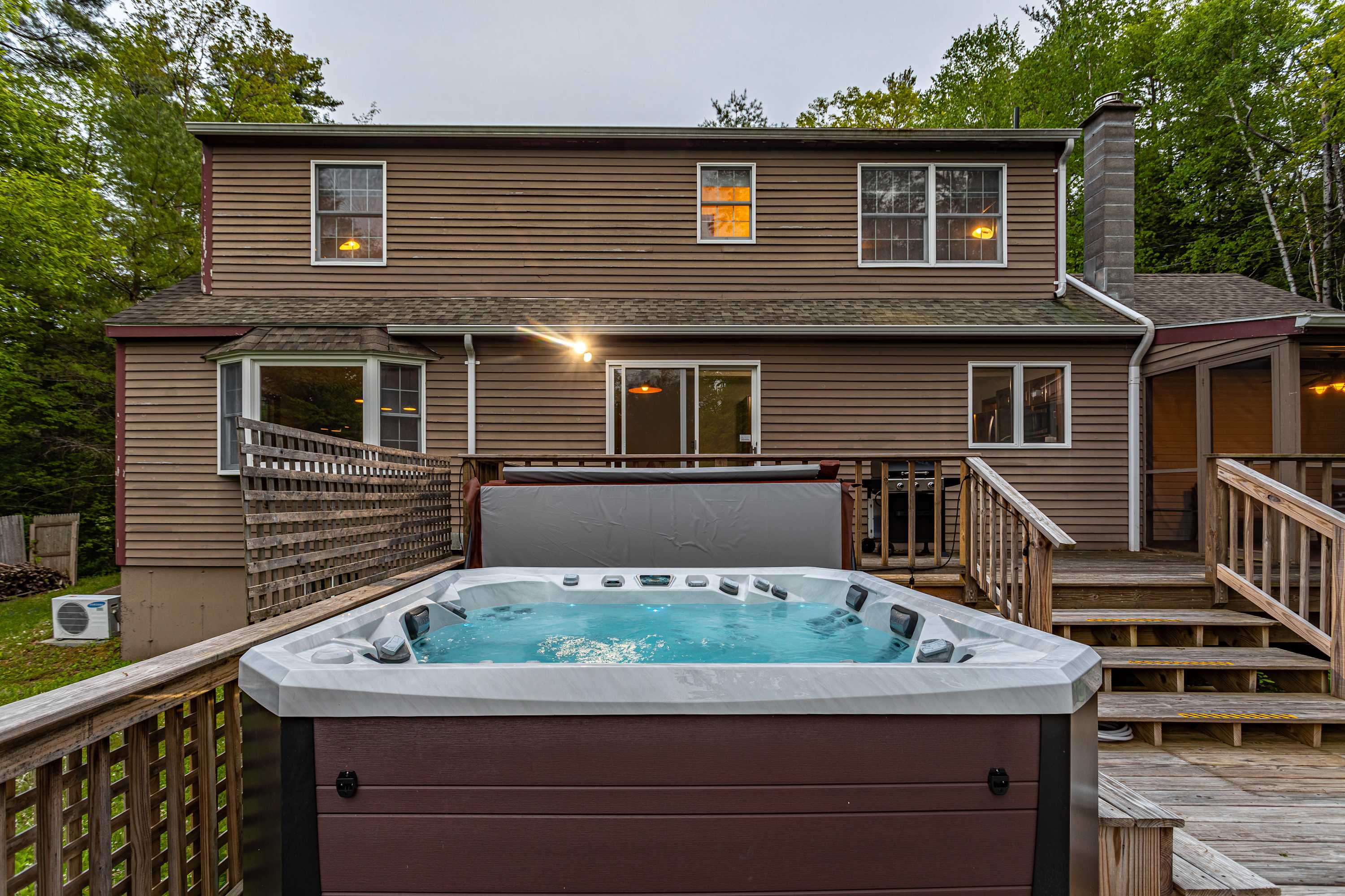A bubbling hot tub with blue water is prominently featured on a wooden deck, with wooden steps leading to the brown-sided house that has illuminated windows and sliding glass doors, all set against a backdrop of green trees.