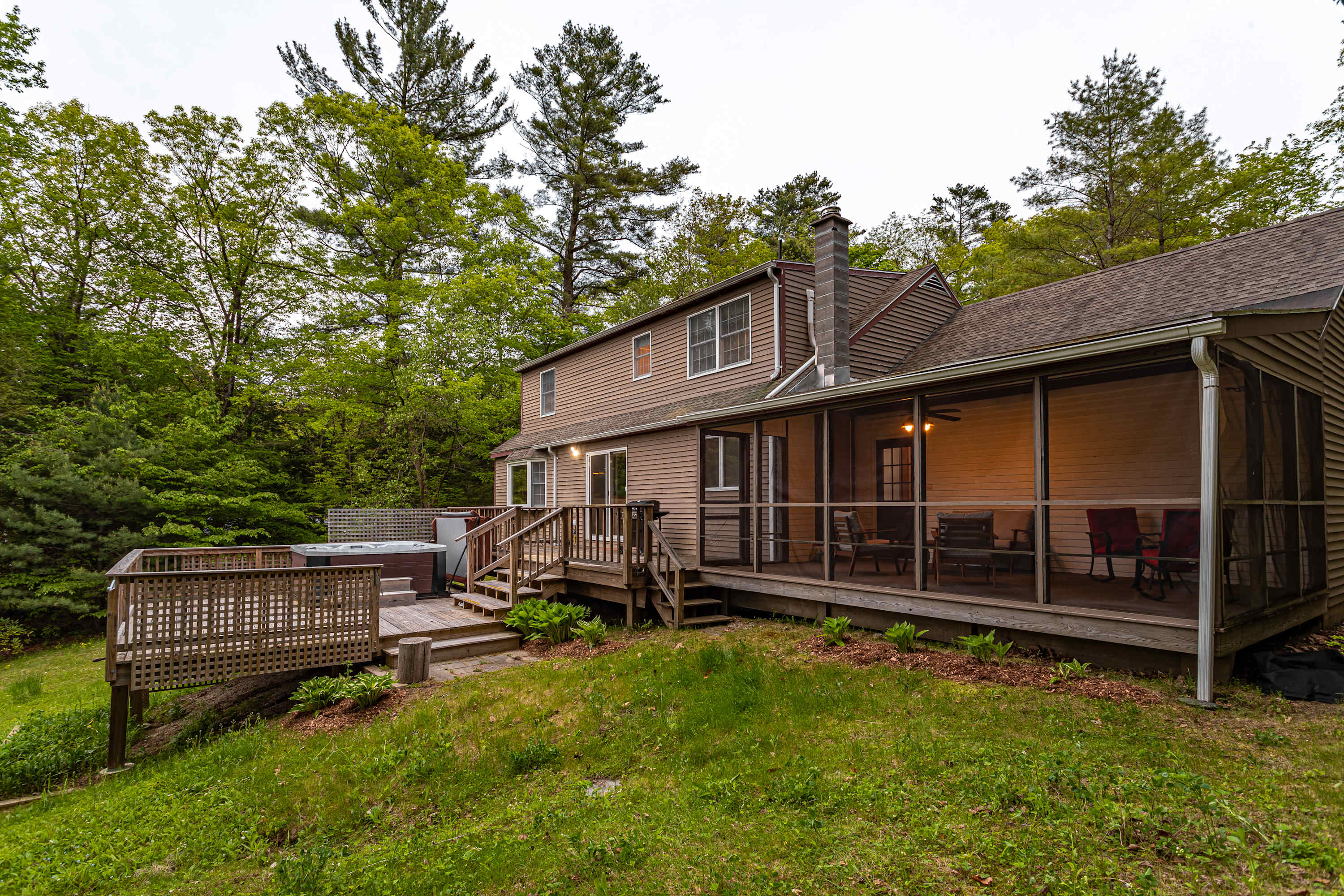 A multi-level wooden deck with a hot tub and a screened-in porch featuring outdoor seating extends from the brown-sided house, surrounded by lush green trees and a grassy slope.
