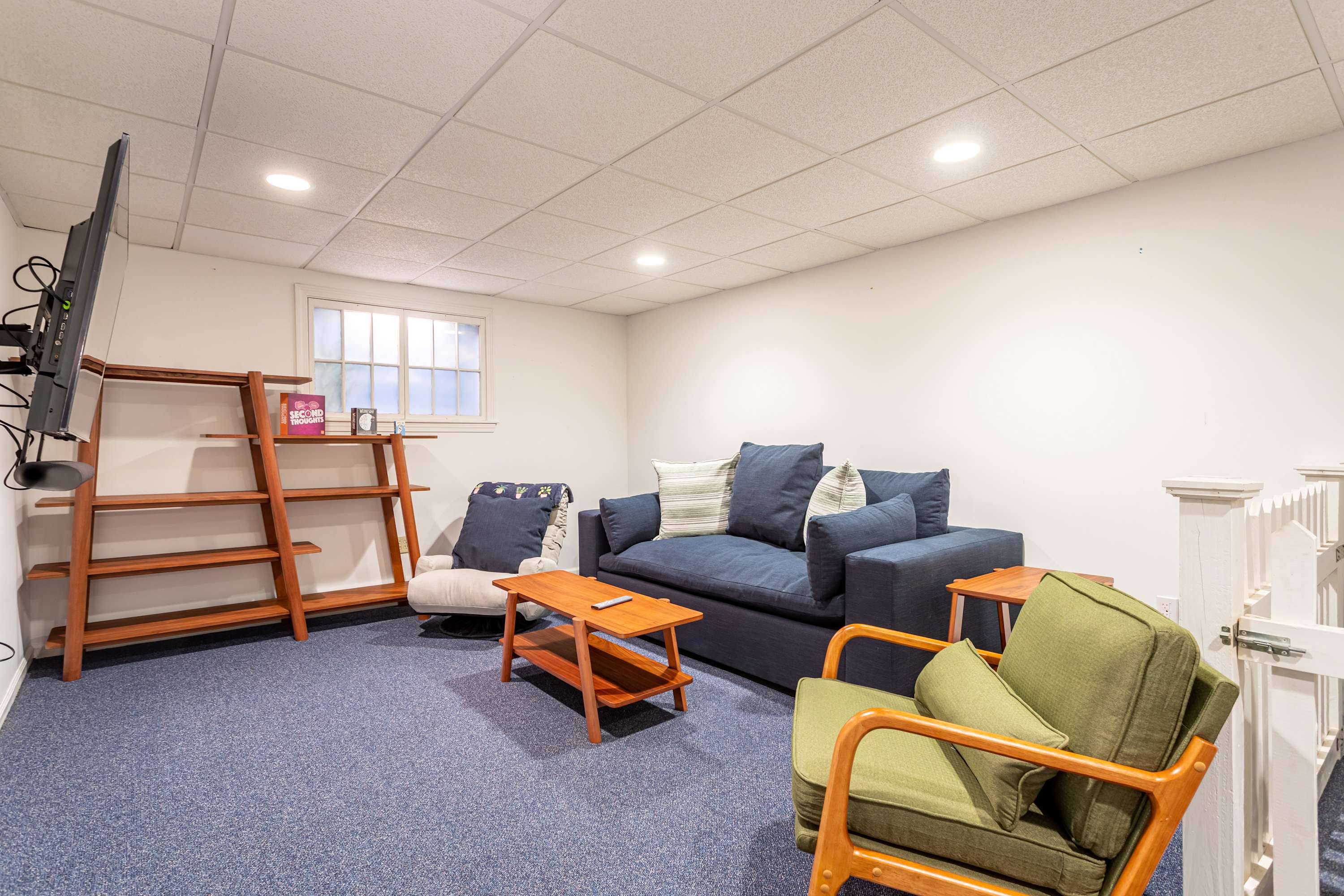 Another perspective of the media room, showing a blue sofa, a green armchair, a wooden bookshelf, and a window, all set on a blue carpet.
