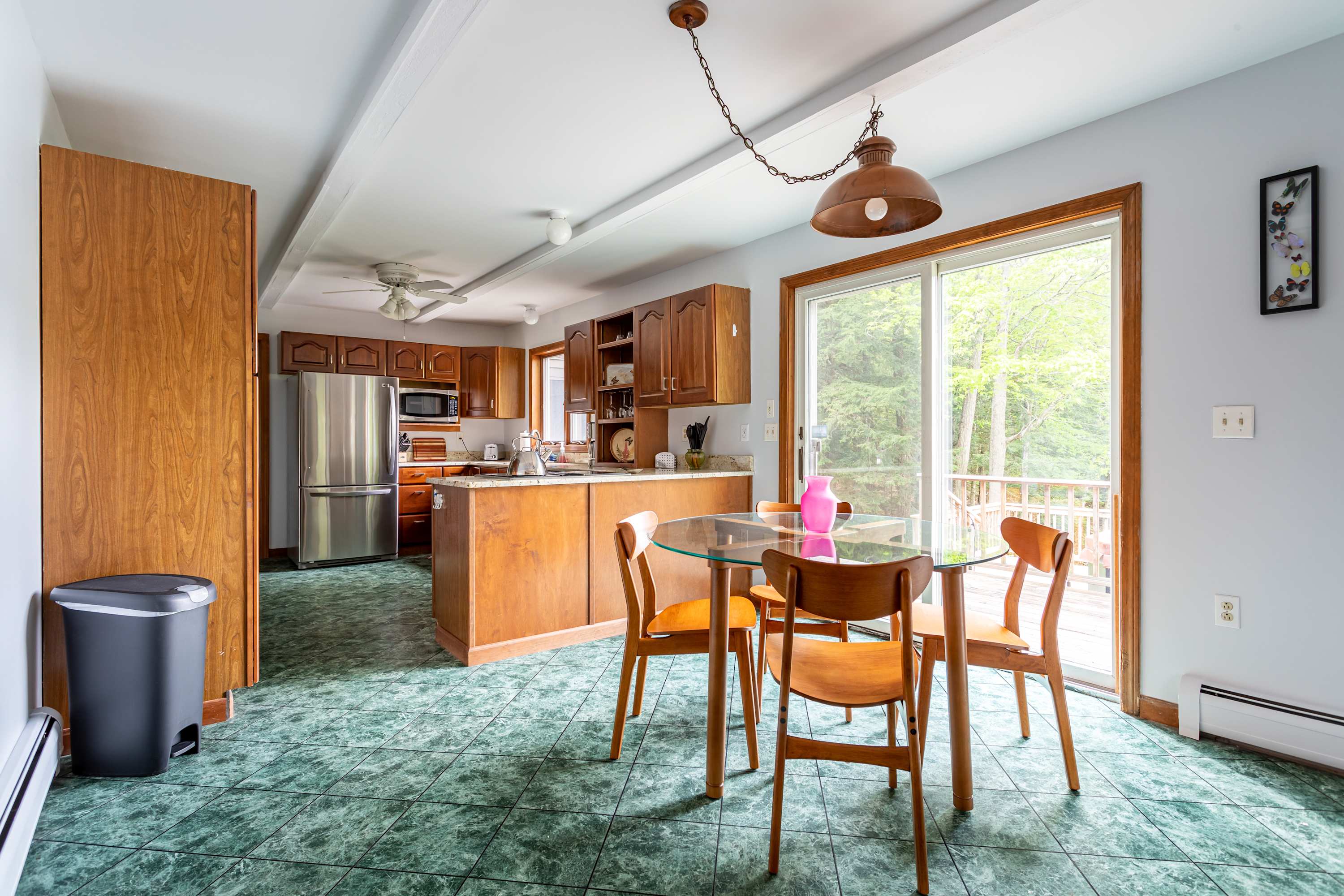 A bright, open-plan kitchen featuring green tiled floors, wooden cabinetry, stainless steel appliances, and a breakfast bar. A glass-top dining table with wooden chairs is adjacent, next to a sliding glass door leading to a deck with outdoor views.