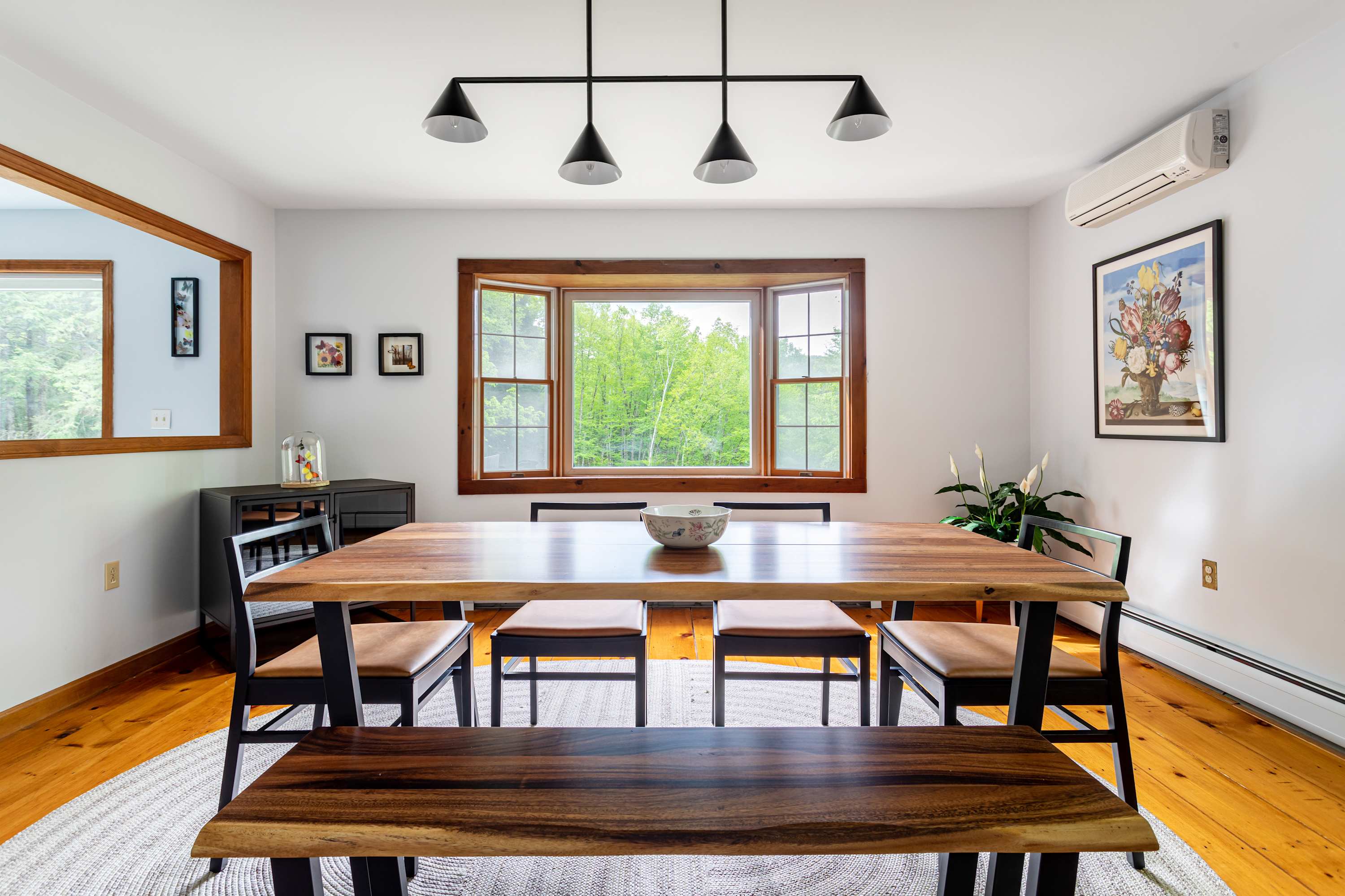A spacious and modern dining room featuring polished hardwood floors and a large live-edge wooden dining table surrounded by black chairs and a matching bench. The room is brightened by a bay window with natural wood trim and illuminated by a sleek black chandelier.