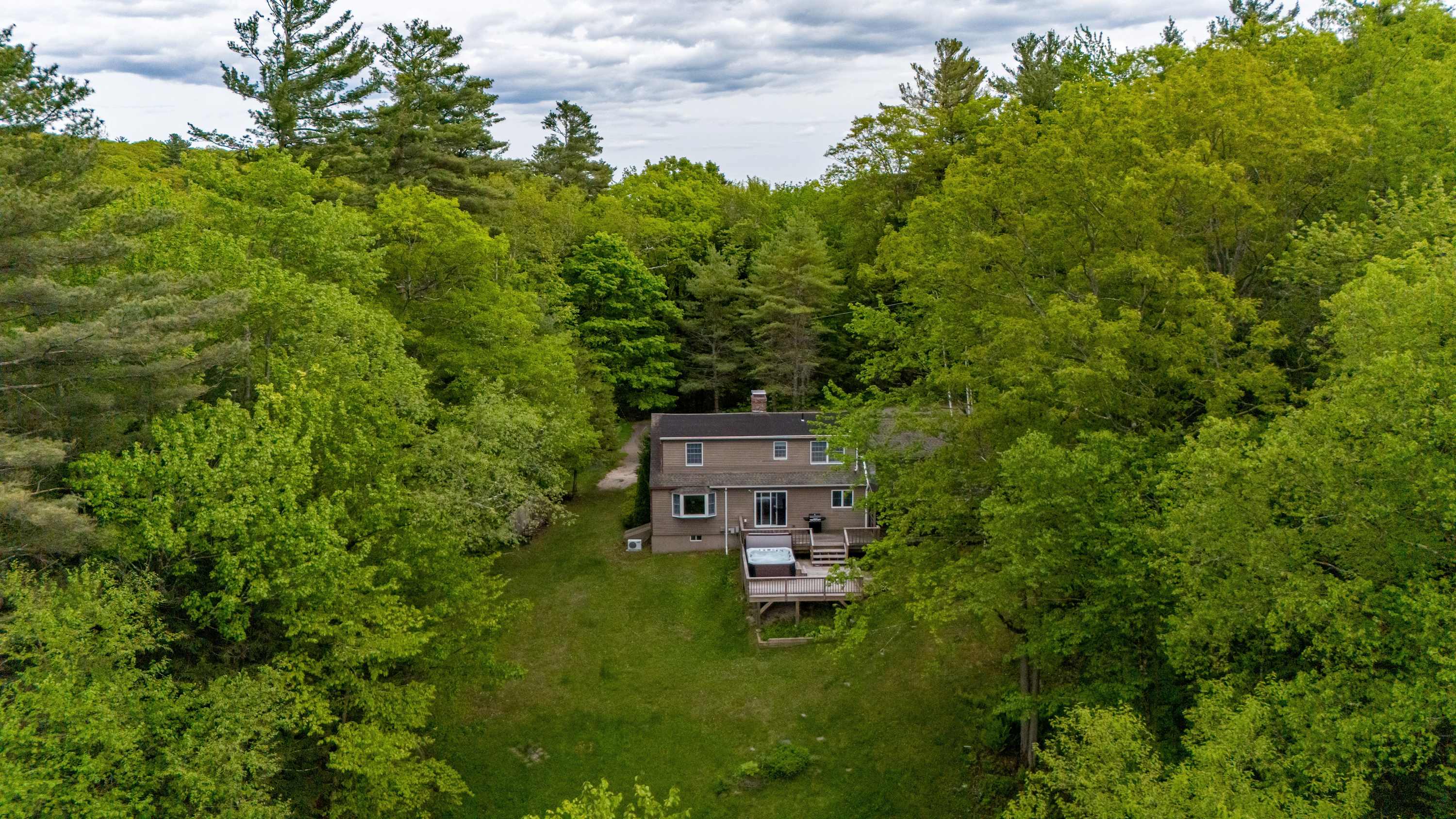 An aerial view showcases a brown-sided house nestled in a clearing amidst a dense, vibrant green forest, with a large wooden deck and hot tub visible at the back of the property.