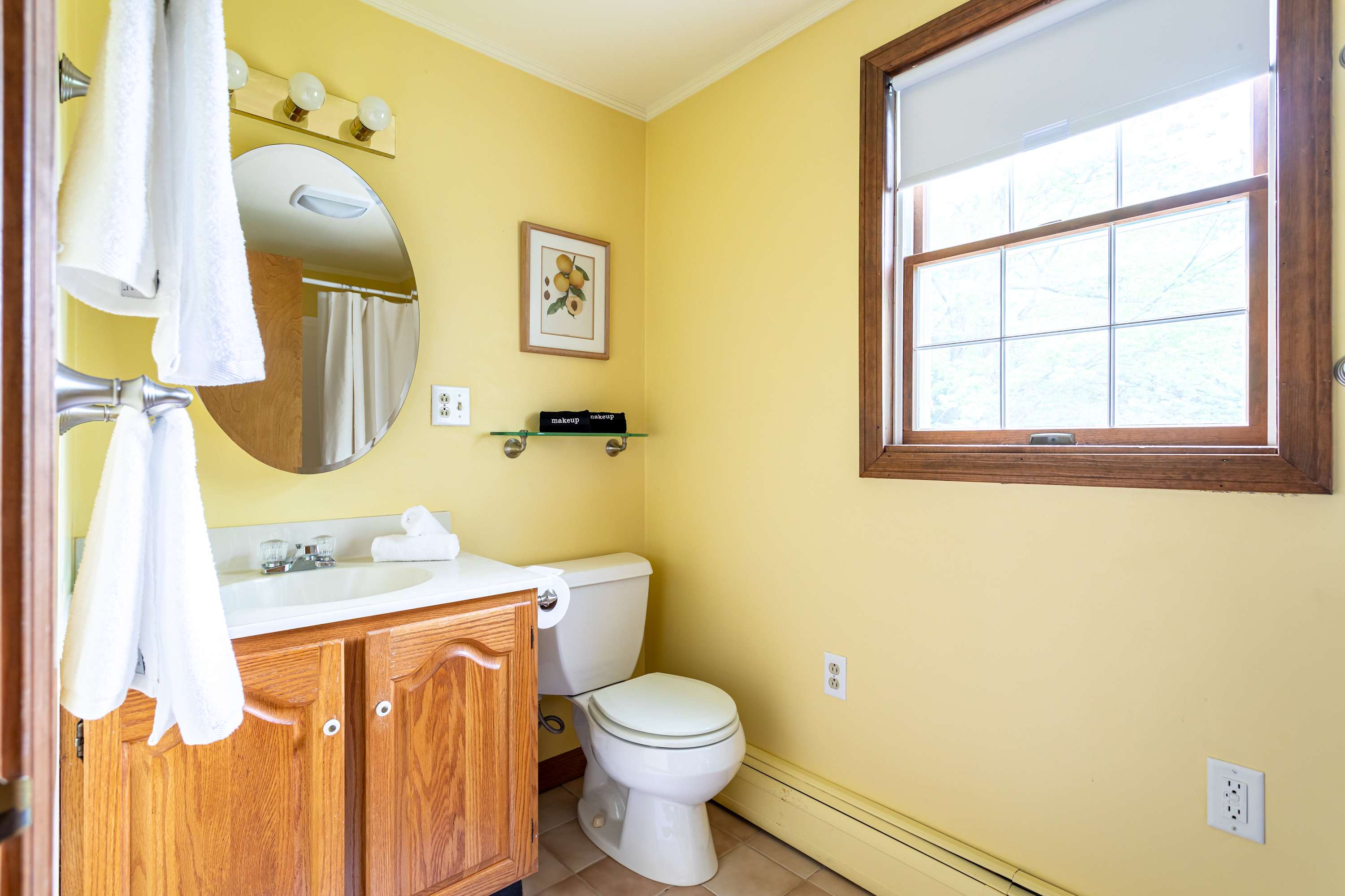 Sunny bathroom with bright yellow walls, a light wood vanity with a white sink, a toilet, and a window with a wooden frame.