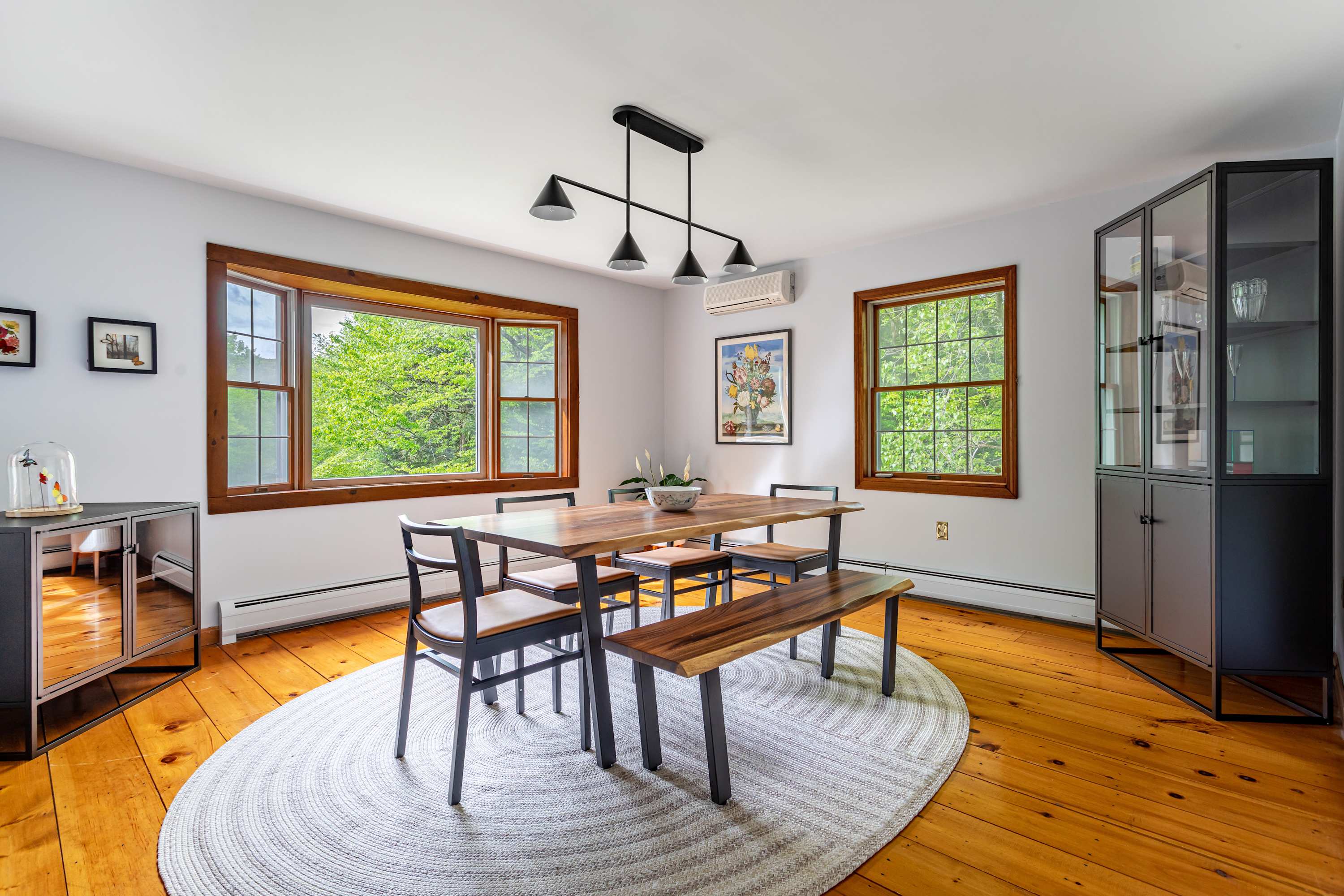 A bright dining room showcasing polished hardwood floors, a large live-edge wooden dining table, and multiple windows with natural wood trim offering views of lush greenery. A modern black chandelier hangs above the table, and a large oval woven rug anchors the space.