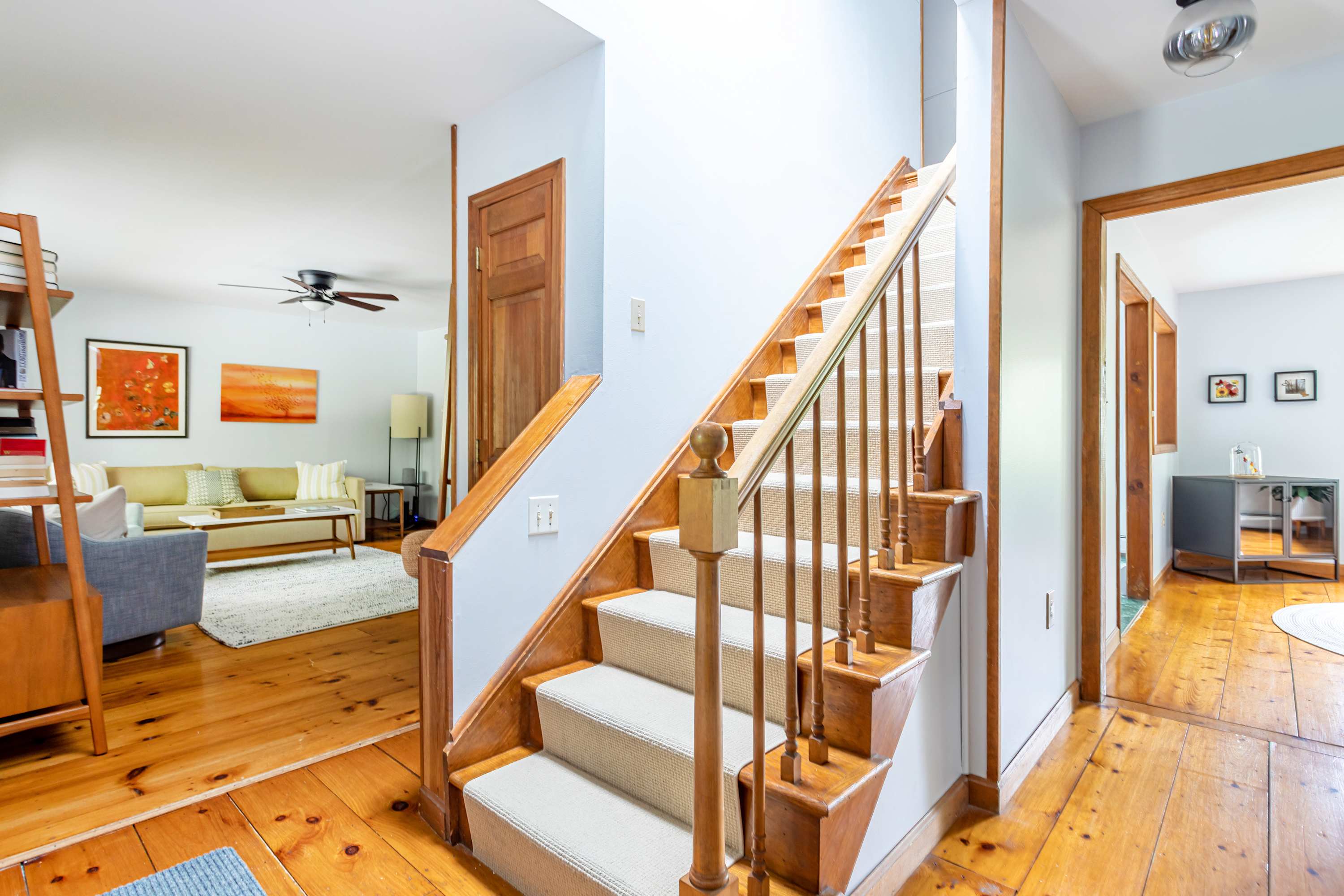 A welcoming foyer featuring polished light wood floors and a carpeted wooden staircase leading to the upper level. An open doorway on the left reveals a living room with a sofa and artwork, creating an inviting transition space.